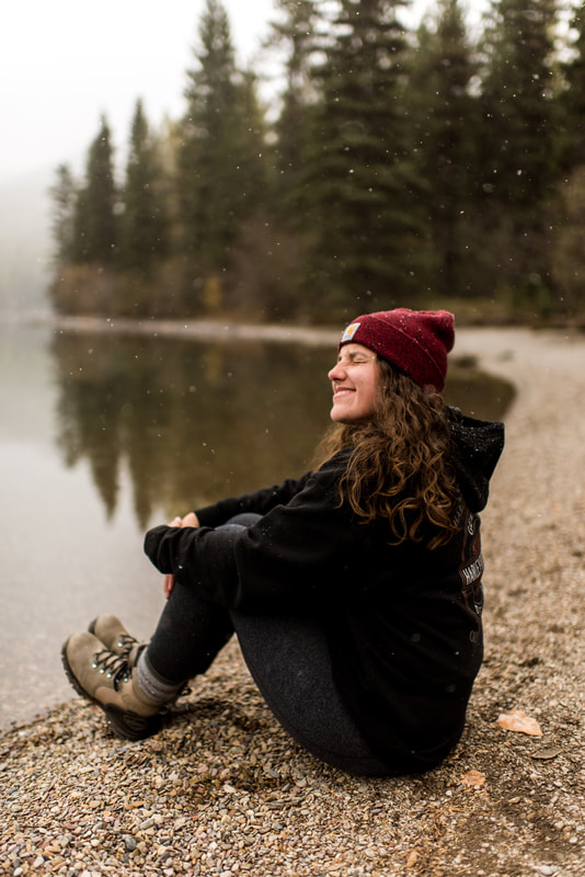 Adventure Elopement Photographer, Jen, sits lakeside in Glacier National Park, Montana while snow falls
