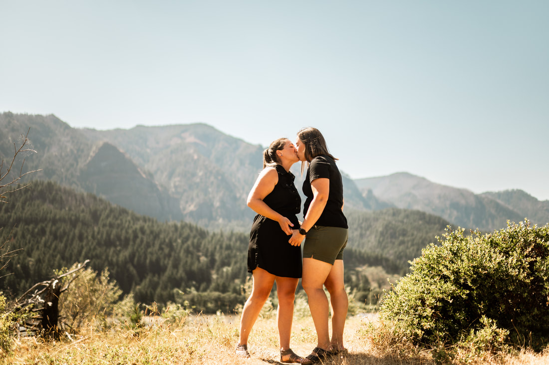 Government Cove Peninsula Engagement Session. Two women hold hands facing each other and kiss in front of mountains.