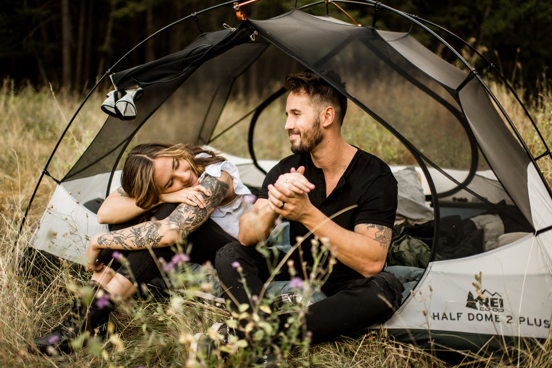 tattooed engaged couple sit in a tent in a golden grass field along Flathead River, Montana outside Glacier National Park as the sun sets